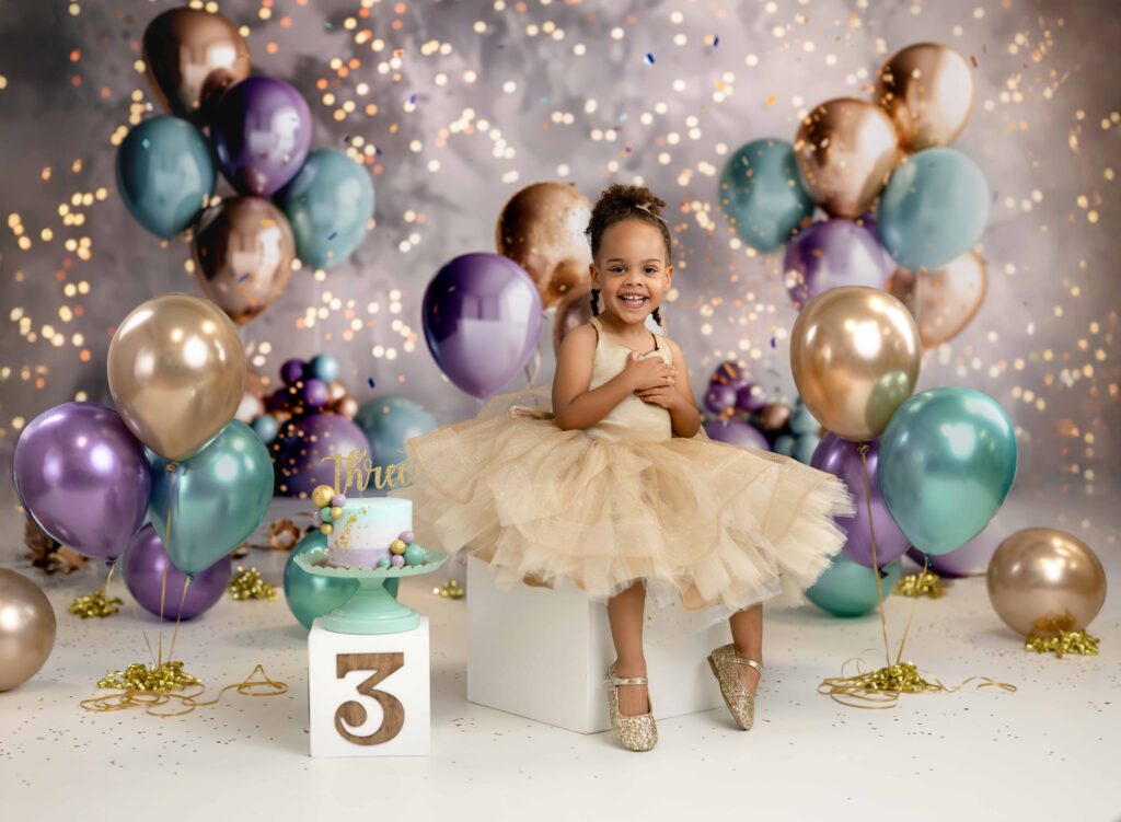 3 year toddler taking birthday photos in a gold dress and smiling. backdrop has teal and gold and lavender balloons.