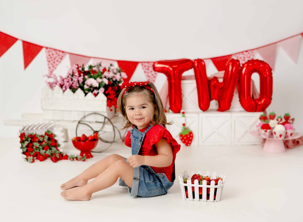 Two sweet strawberry birthday set with 2 year old girl in overalls and red shirt smiling.