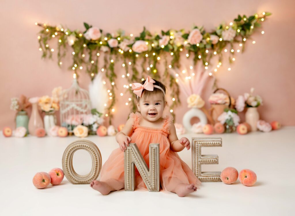 Baby girl in peach dress smiling with ONE letters by her.