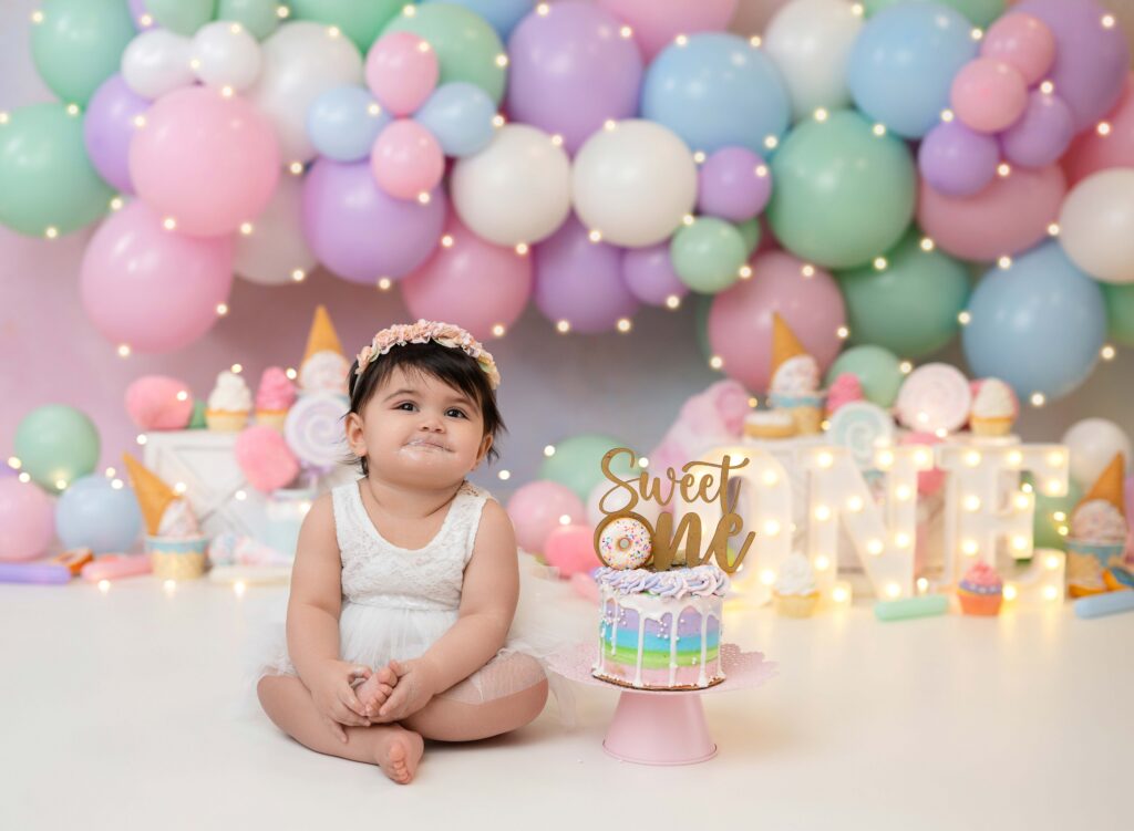 Baby girl sitting next to sweet one cake and grinning. 