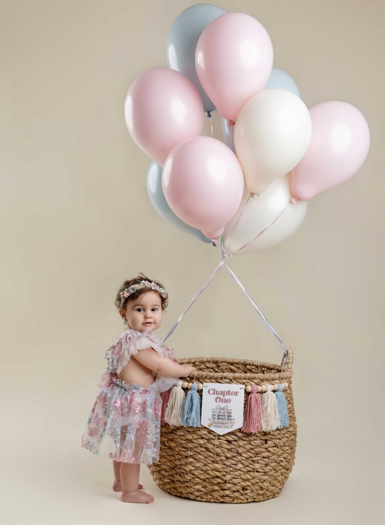 Baby standing next to basket of balloons with 1st chapter banner on it.