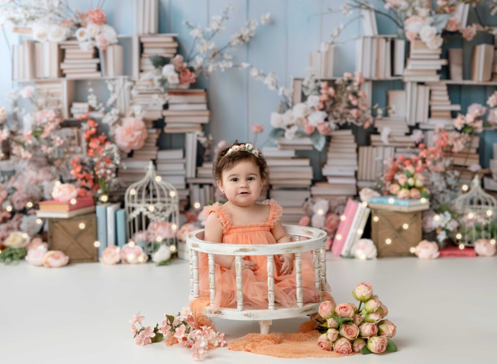 Baby girl in peach dress, sitting in a white wooden crate with florals around her.