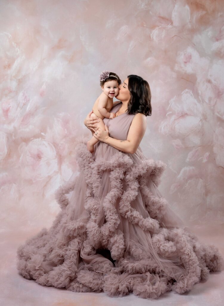 Mom in mauve gown holding naked baby and kissing her cheek.