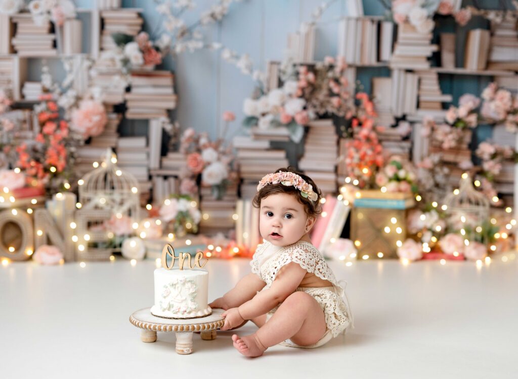 Baby girl reaching for birthday cake for her 1st birthday photos on a Chapter One birthday theme backdrop.