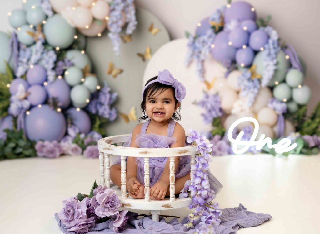 Baby girl in purple dress sitting in white rounded crate smiling.