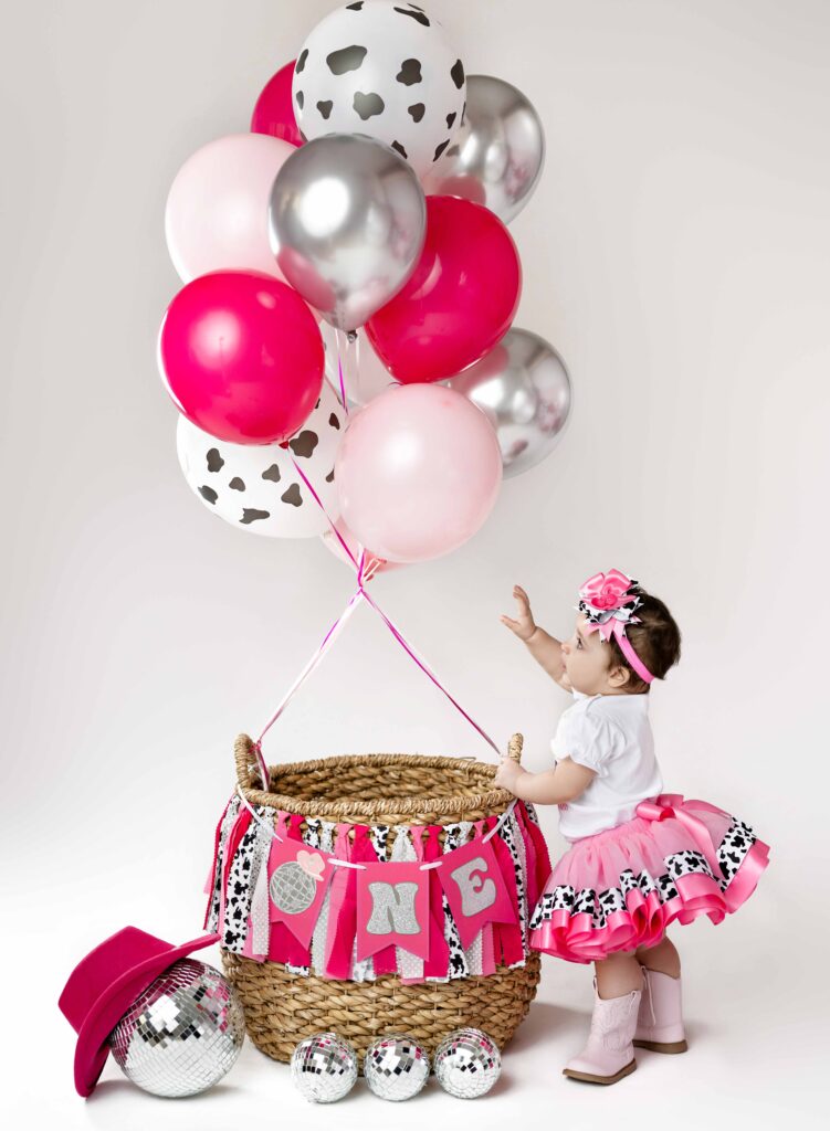 Baby girl holding onto basket and reaching for balloons. 1st Birthday photo taken at Atlanta photo studio.