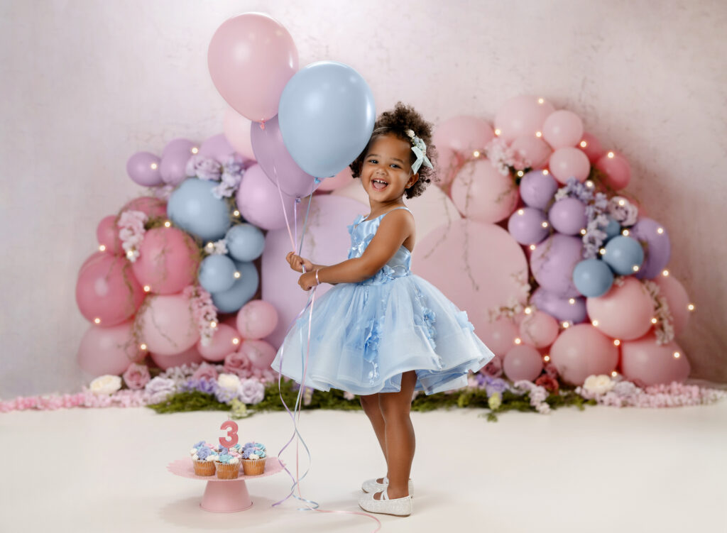 3 year girl i blue dress, holding pastel balloons and smiling for her 3rd birthday photoshoot in Atlanta.