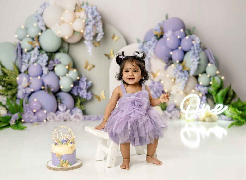 Baby girl sitting on stool smiling in front of a purple and sage green balloon garland birthday set.