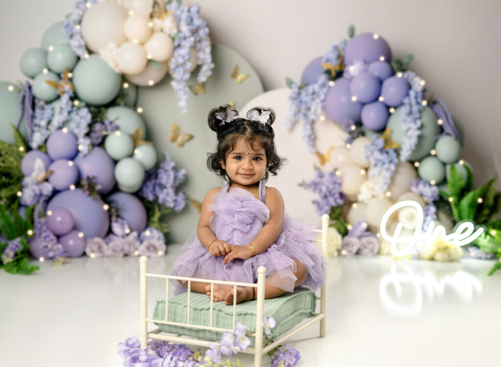 Baby girl in purple dress sitting on tiny bed prop, taking 1st birthday photos.