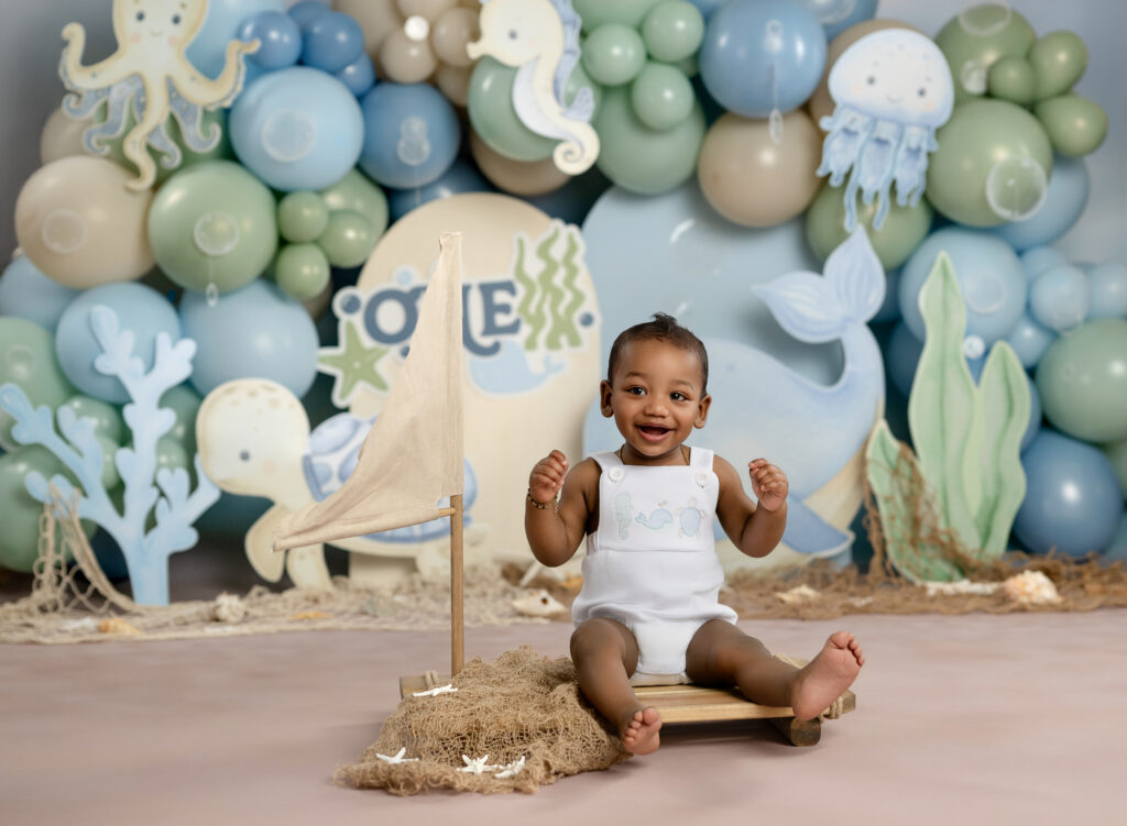 baby boy sitting on a sail boat smiling in front of an under the sea cake smash set.