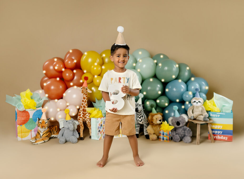 Toddler boy standing in front of balloon garland and stuffed animals with party hats.
