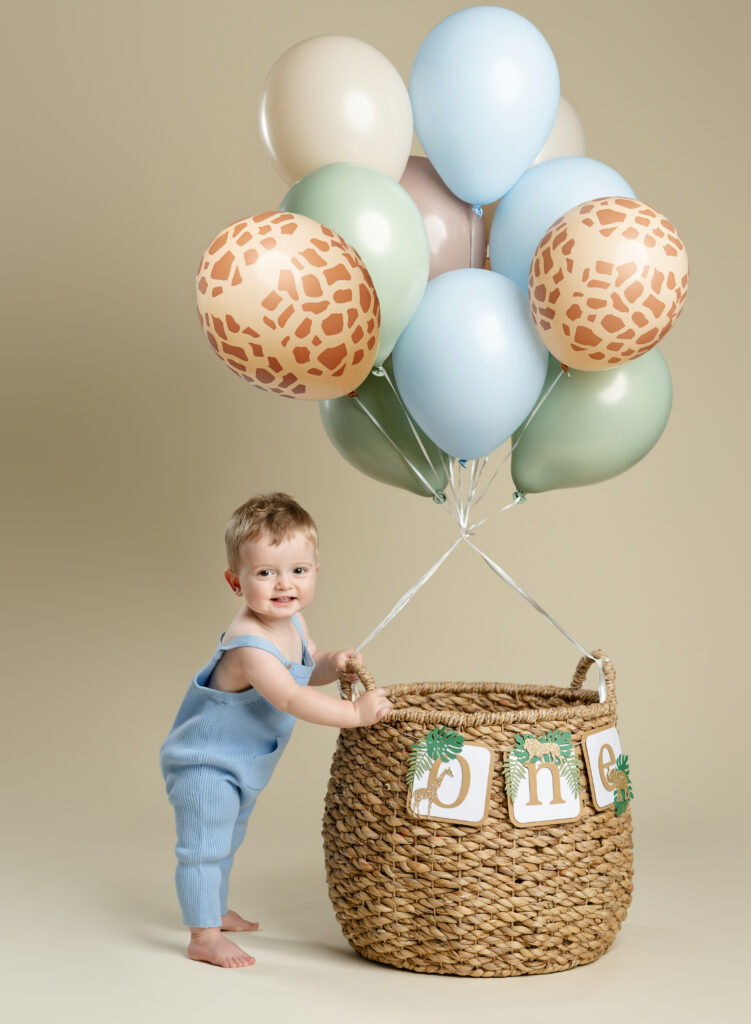 Baby boy taking birthday photos next to a safari balloon basket prop.