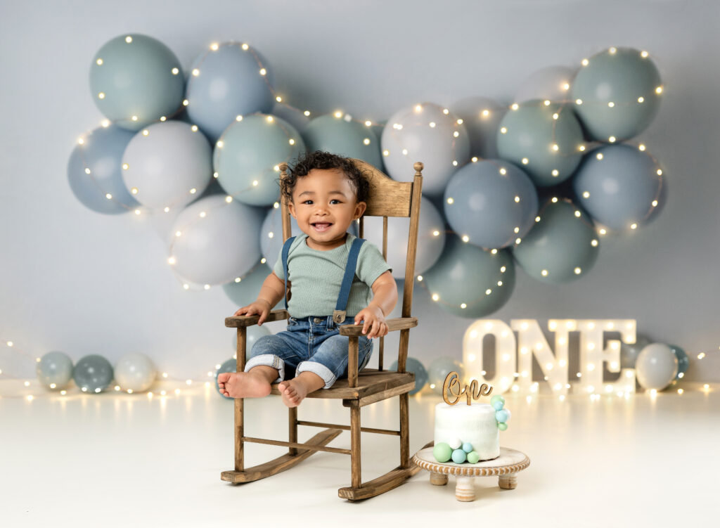 Baby boy sitting in wooden rocking chair in front of balloon garland for 1st birthday photoshoot.