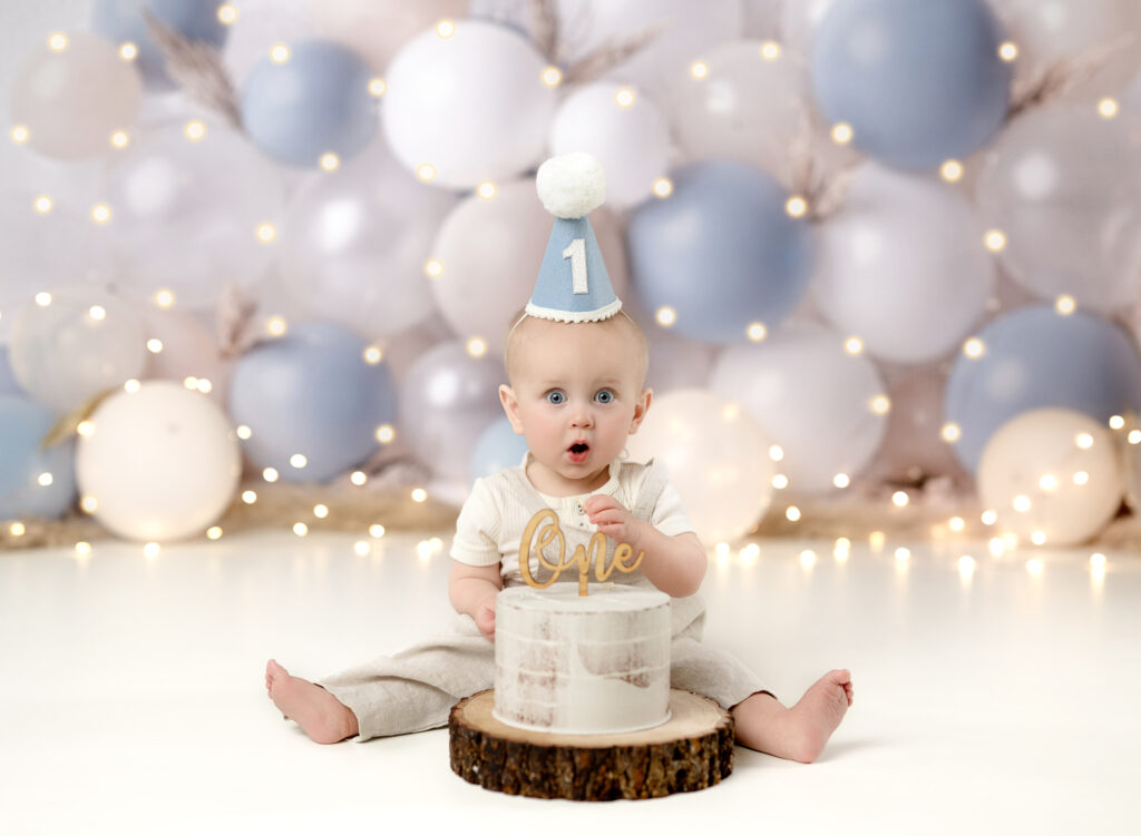 Baby boy eating cake for his 1st birthday photos, in front of a white, cream and blue balloon garland backdrop.