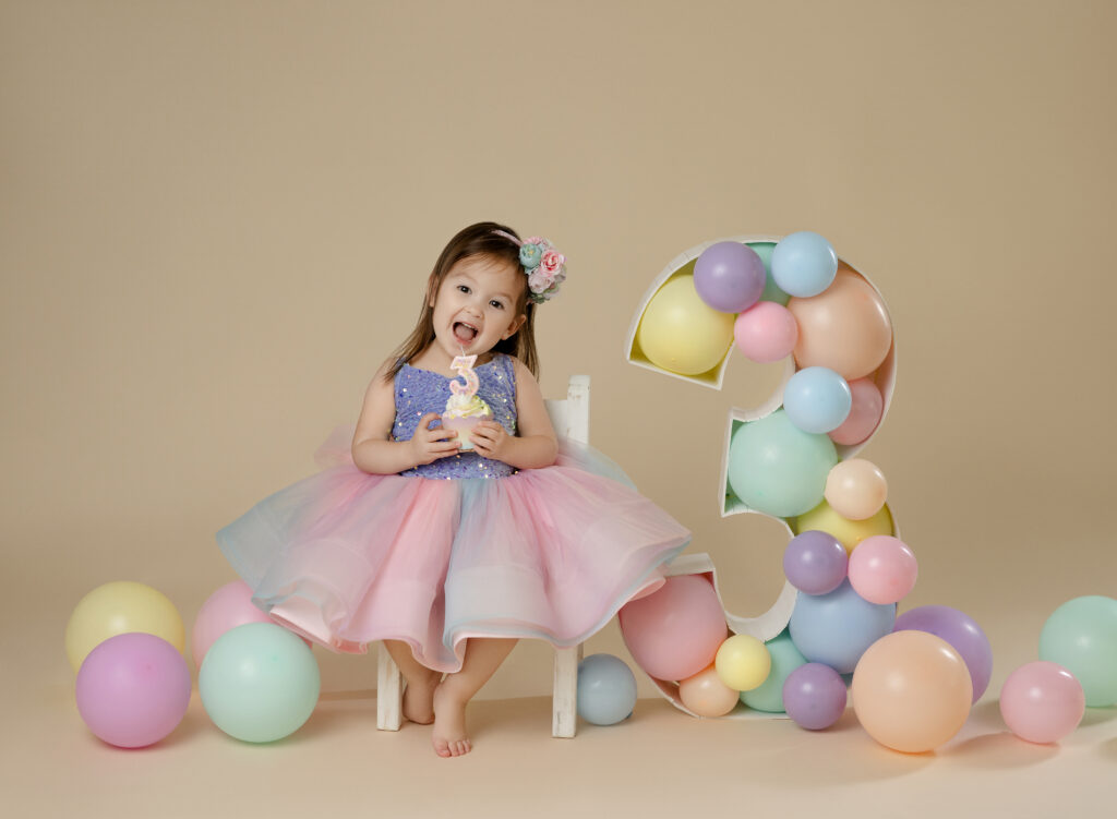 toddler girl sitting next to balloon filled marquee with a cupcake and big surprise on her face.