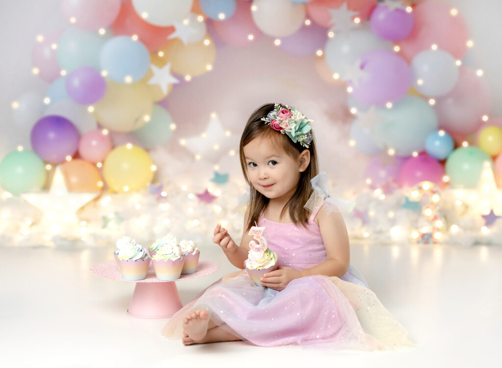 3 year old birthday girl eating cupcakes in front of a pastel balloon garland