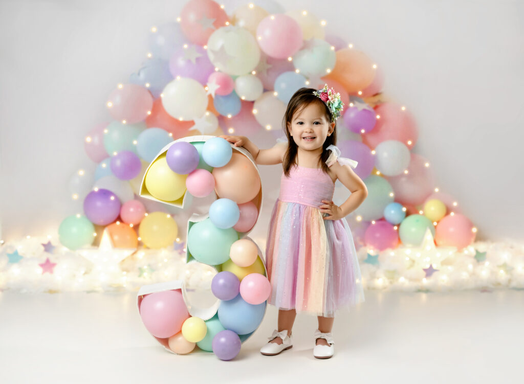 3 year old birthday girl standing by a #3 balloon filled marquee smiling in front of a pastel balloon garland