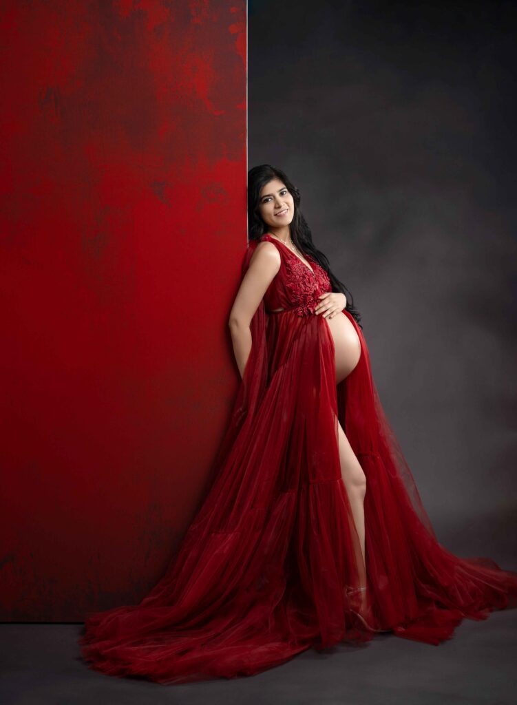 Pregnant women in red dress leaning on a tall red panel backdrop and holding her baby bump.