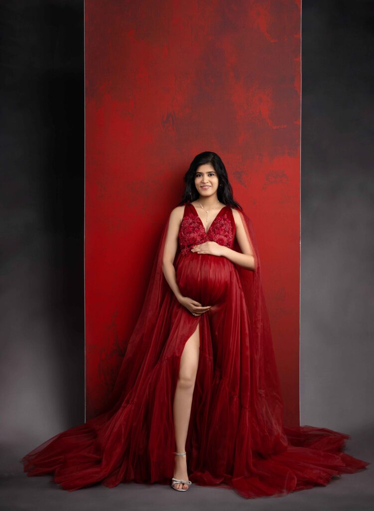Pregnant Mom in red maternity dress, holding her belly and looking at the camera smiling. 