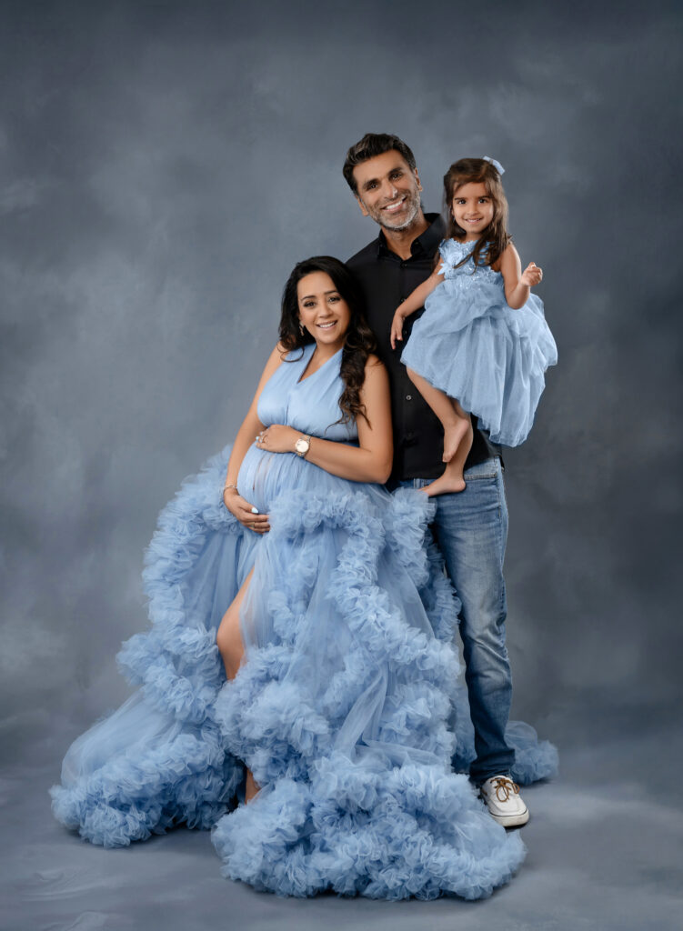 Dad holding toddler girl and Mom in blue maternity dress holding her baby bump. Everyone smiling at the camera.