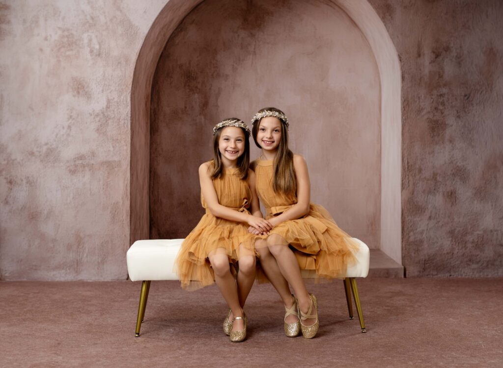 2 sisters sitting on a bench in front of tuscan arch and smiling at camera. 