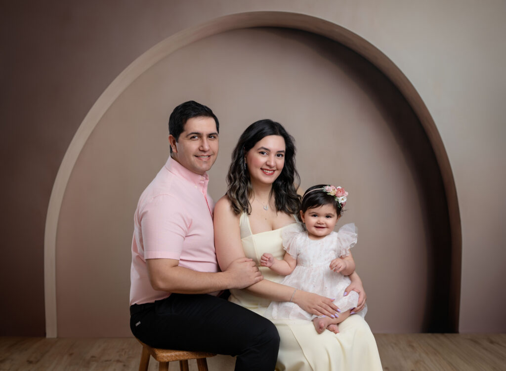 Family of 3 sitting in front of an arch backdrop and smiling at the camera.
