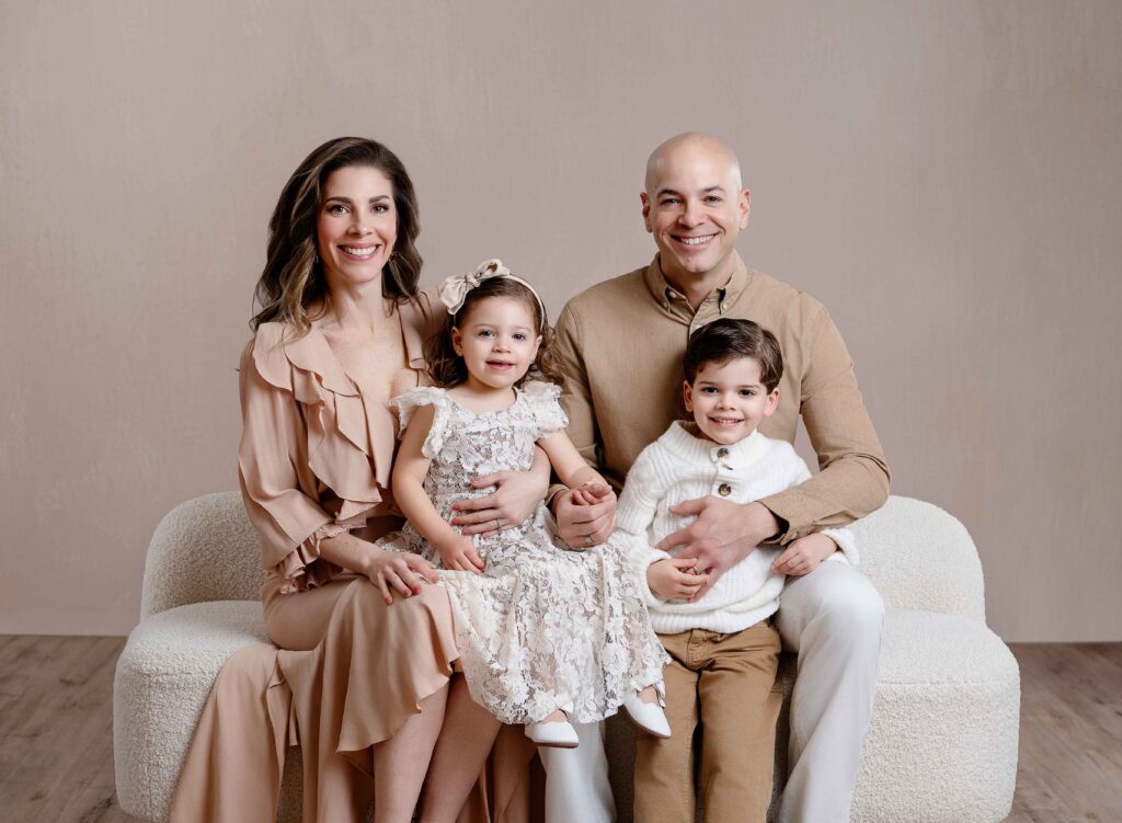 Family of 4 sitting on couch in front of a cream wall and smiling at camera.