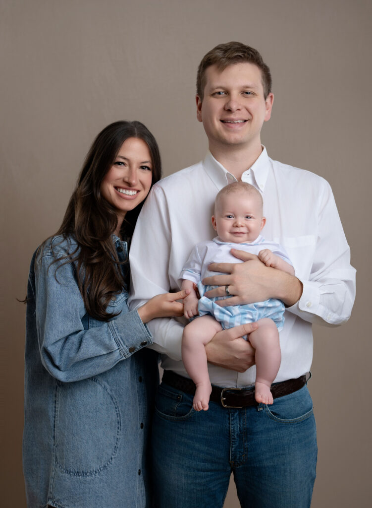 Dad holding infant baby boy and Mom holding his hand and smiling at the camera.