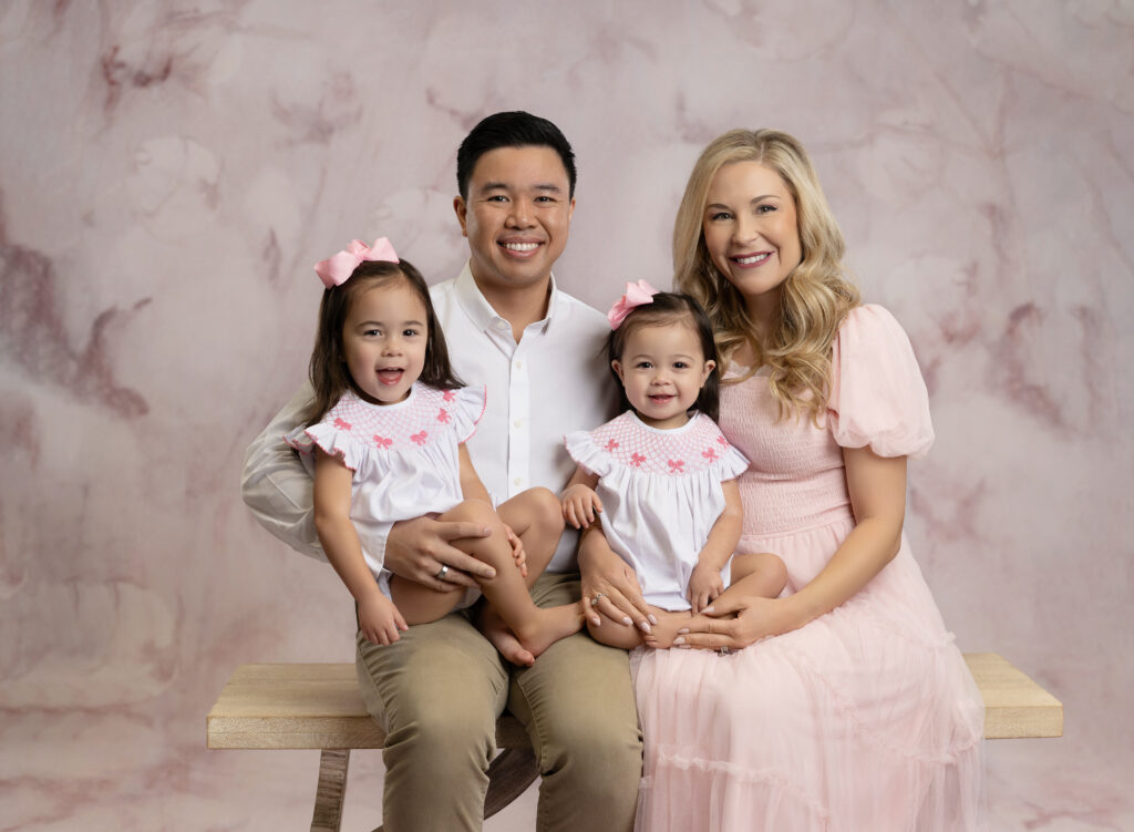 family in pink and white clothes taking studio photo on a bench.