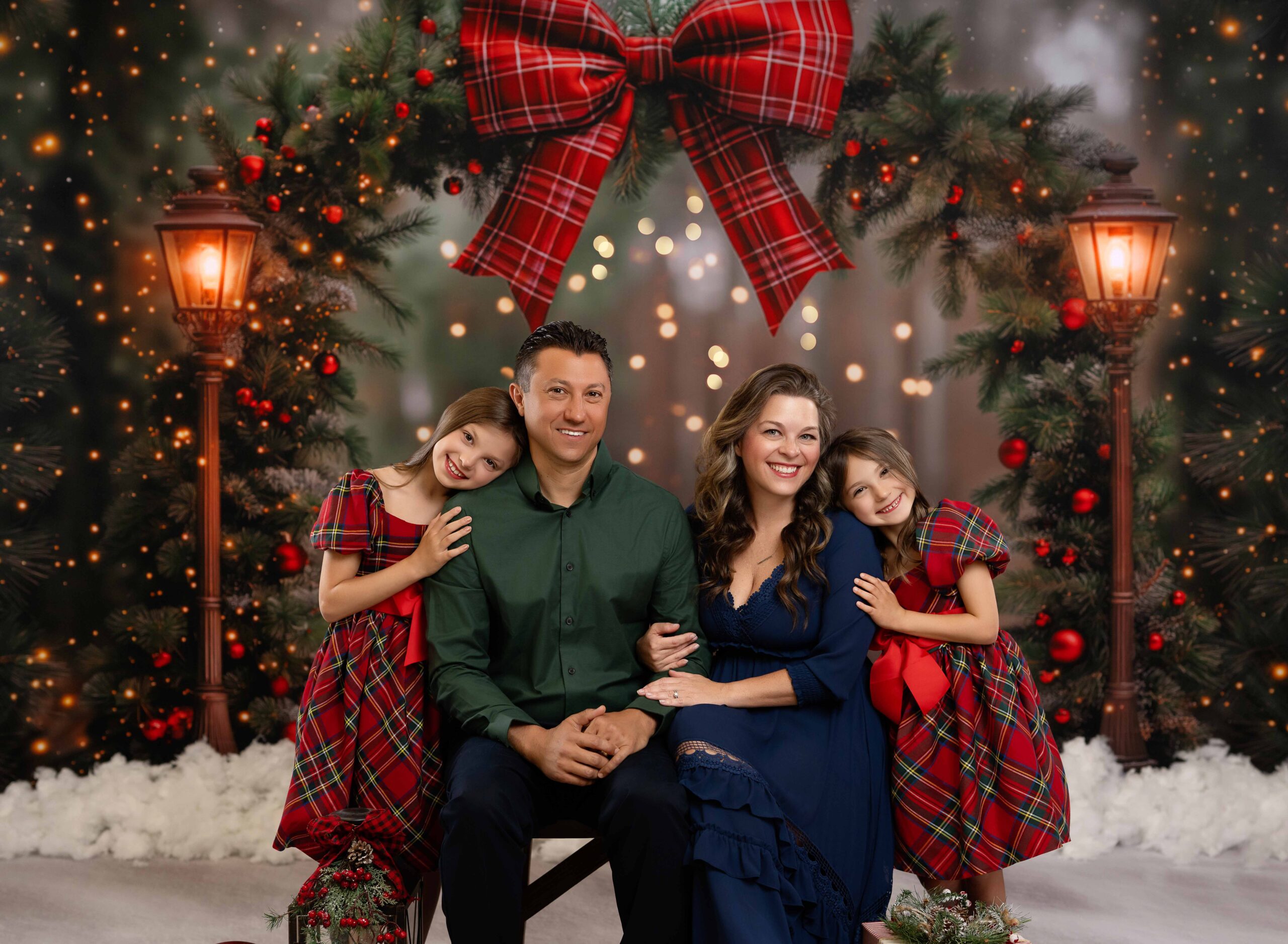 Mom and Dad sitting on a bench with girls in plaid dresses on each side smiling. They are being photographed on a Christmas arch backdrop by Atlanta family photographer- Jenni Nikki Photography.