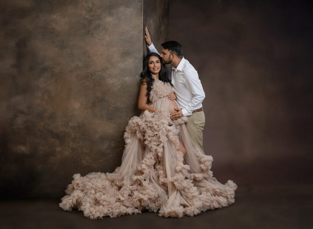 Couple taking maternity photos in studio on brown backdrop.