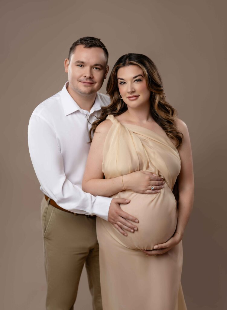 Mom wrapped in cream drape with Dad hugging her and both holding the baby bump and looking at camera with soft expressions.