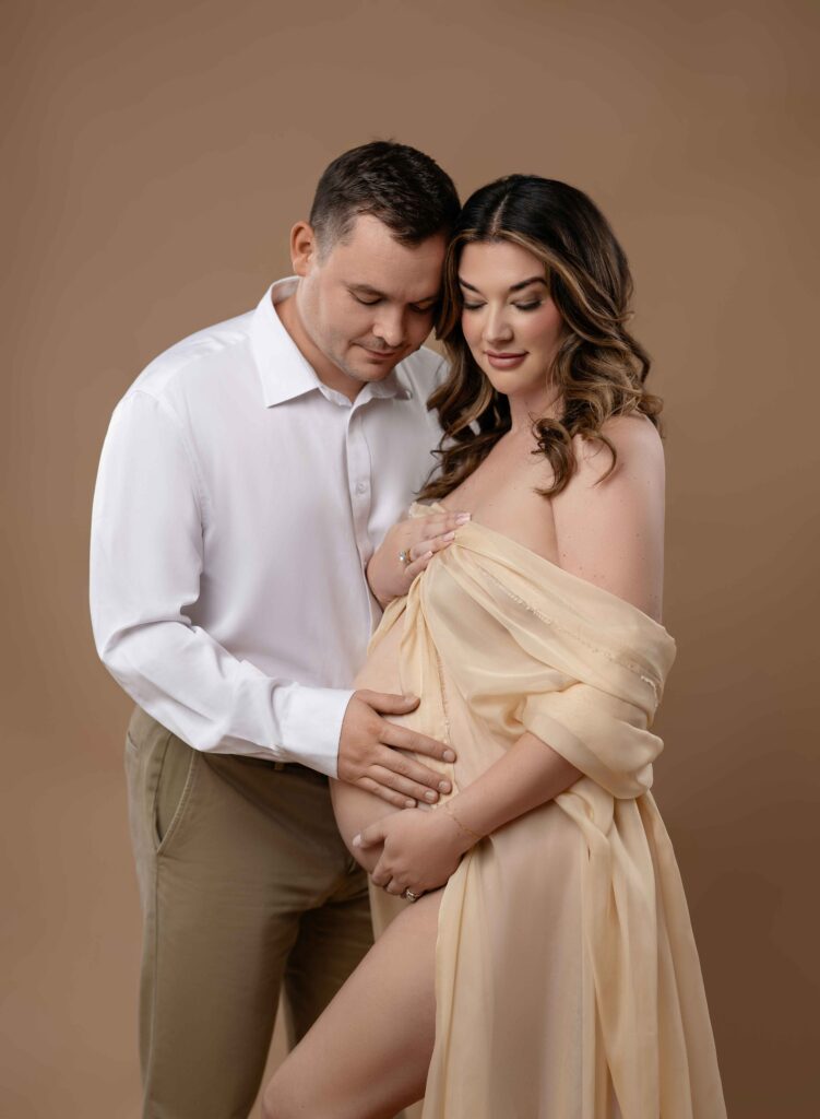 Mom in cream drape looking at belly and Dad snuggling in holding the belly as well.