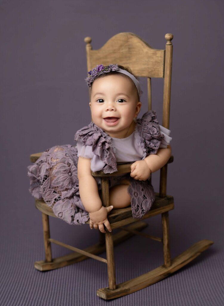Baby girl in purple romper sitting in wooden rocking chair.