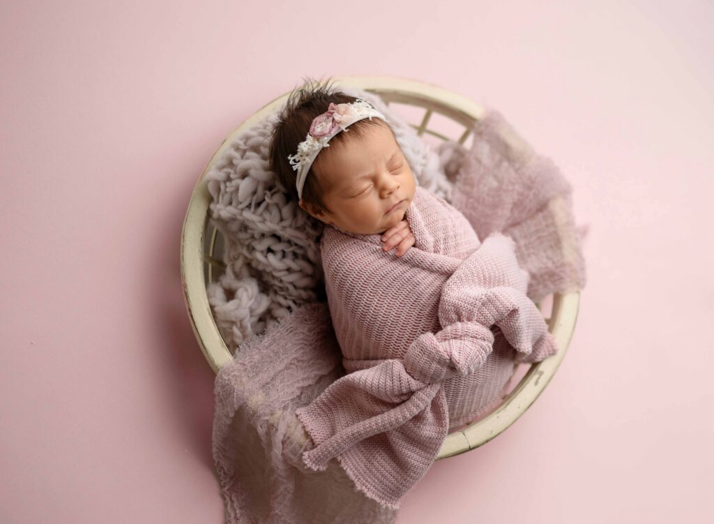Baby swaddled in pink wrap and sleeping in a cream bowl.