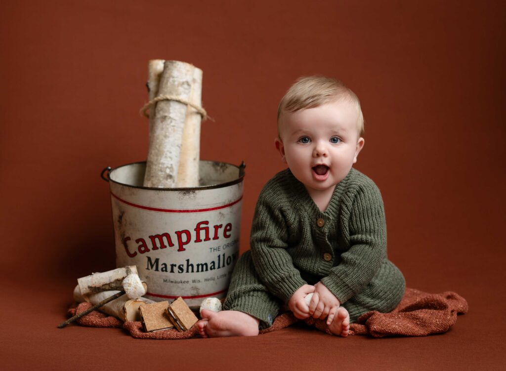 Baby sitting next to campfire bucket with wood and smores.