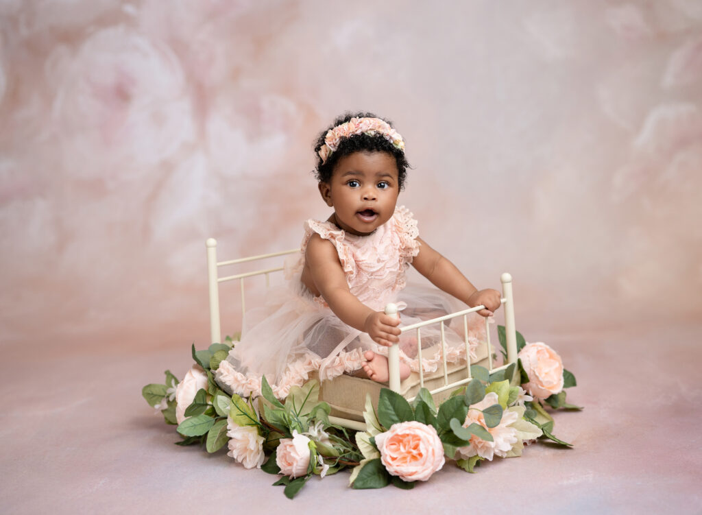 Baby girl sitting in metal cream bed with flowers around it.