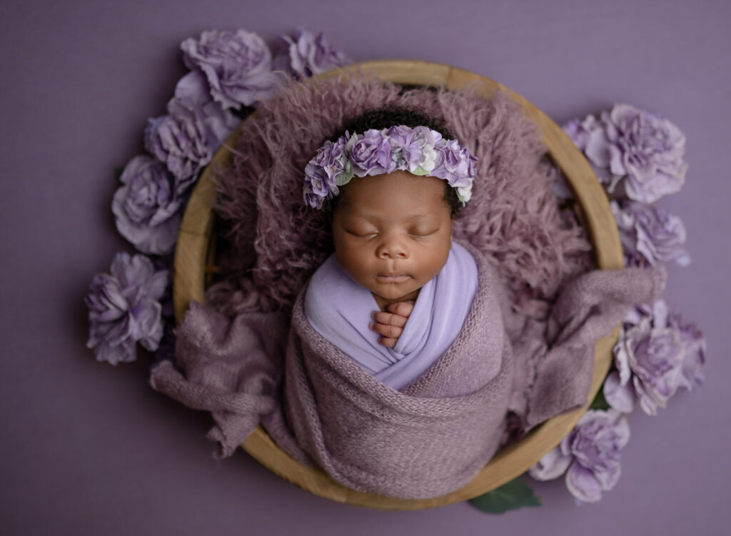 Newborn baby girl in lavender swaddle sleeping in a bowl surround by purple florals.