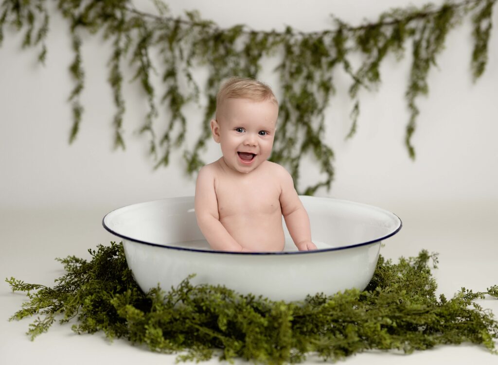 Baby boy in tub with milk bath and greenery smiling at camera. Milk bath mini sessions were done at Woodstock-Atlanta baaby photo studio.