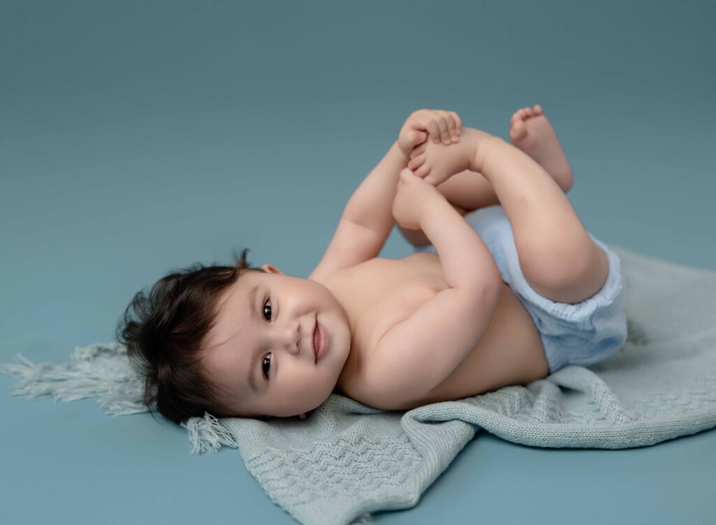 Baby in a blue diaper cover grabbing his toes and smiling. Photographed at Atlanta baby photo studio in Woodstock, GA.