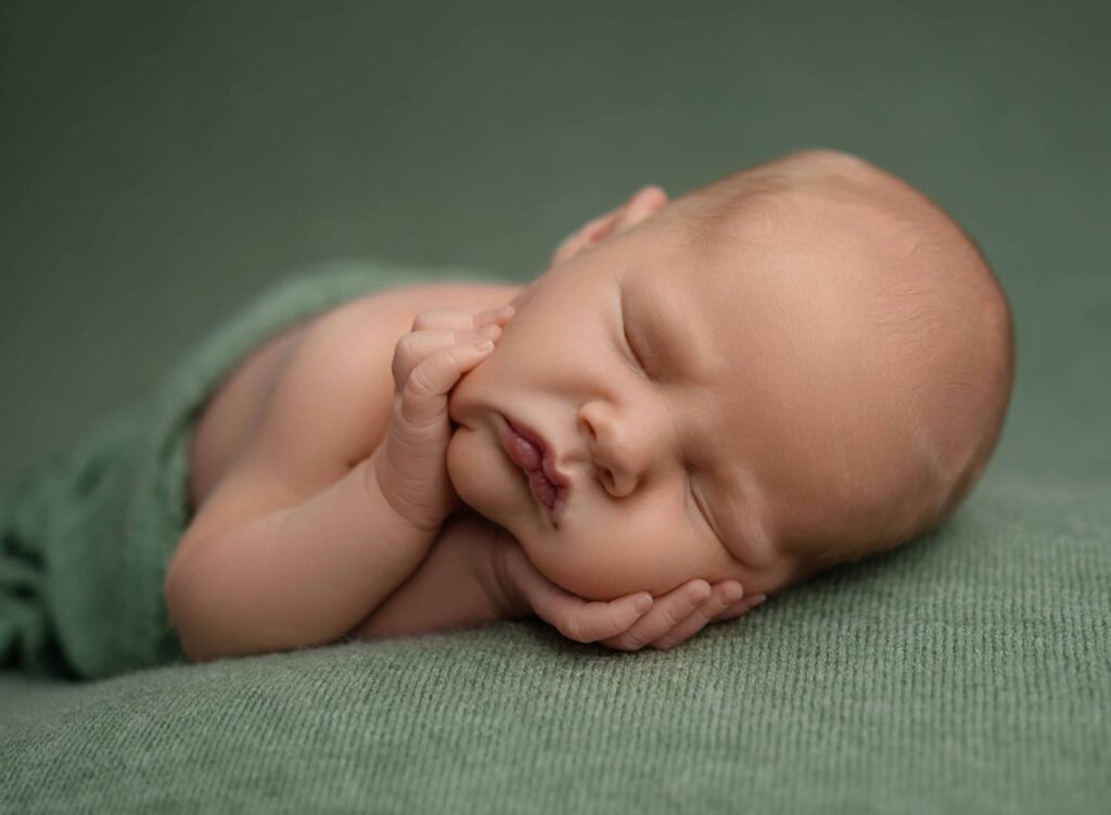 Baby boy sleeping with hands on cheeks and green sage backdrop