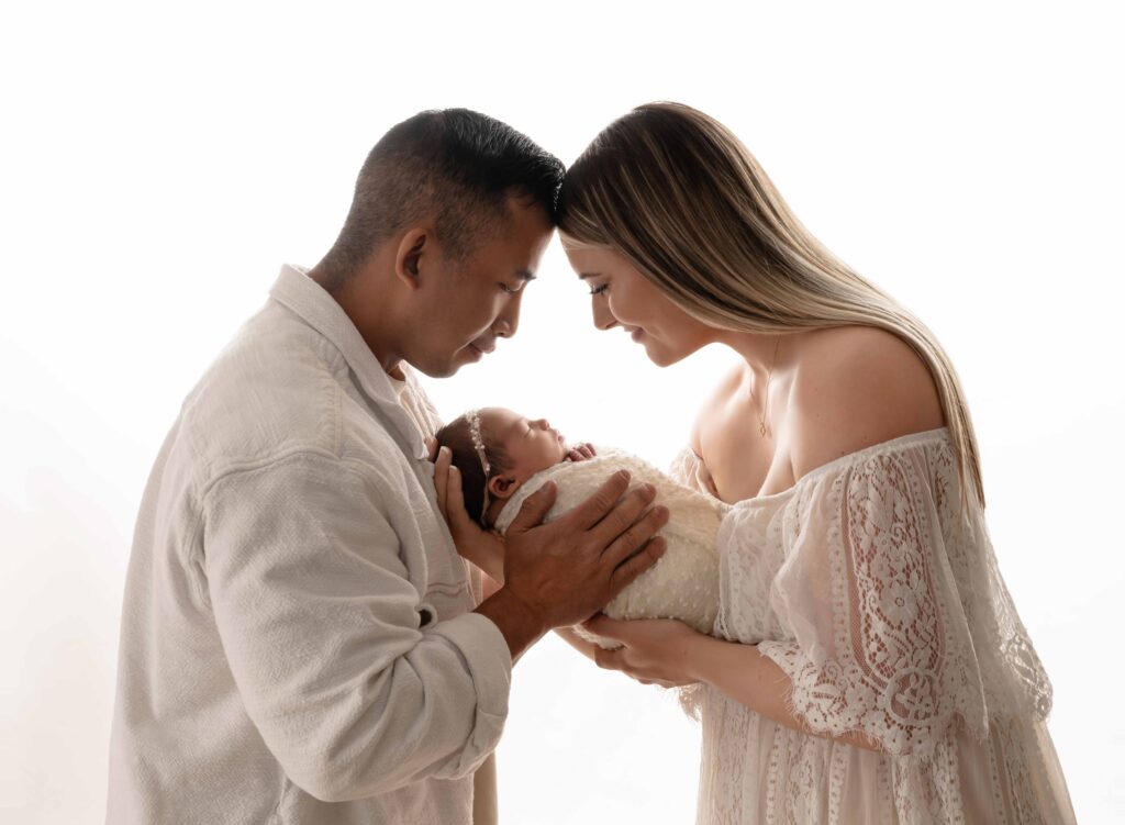 Profile photo of Mom and Dad smiling at baby girl with a white backlighting backdrop.