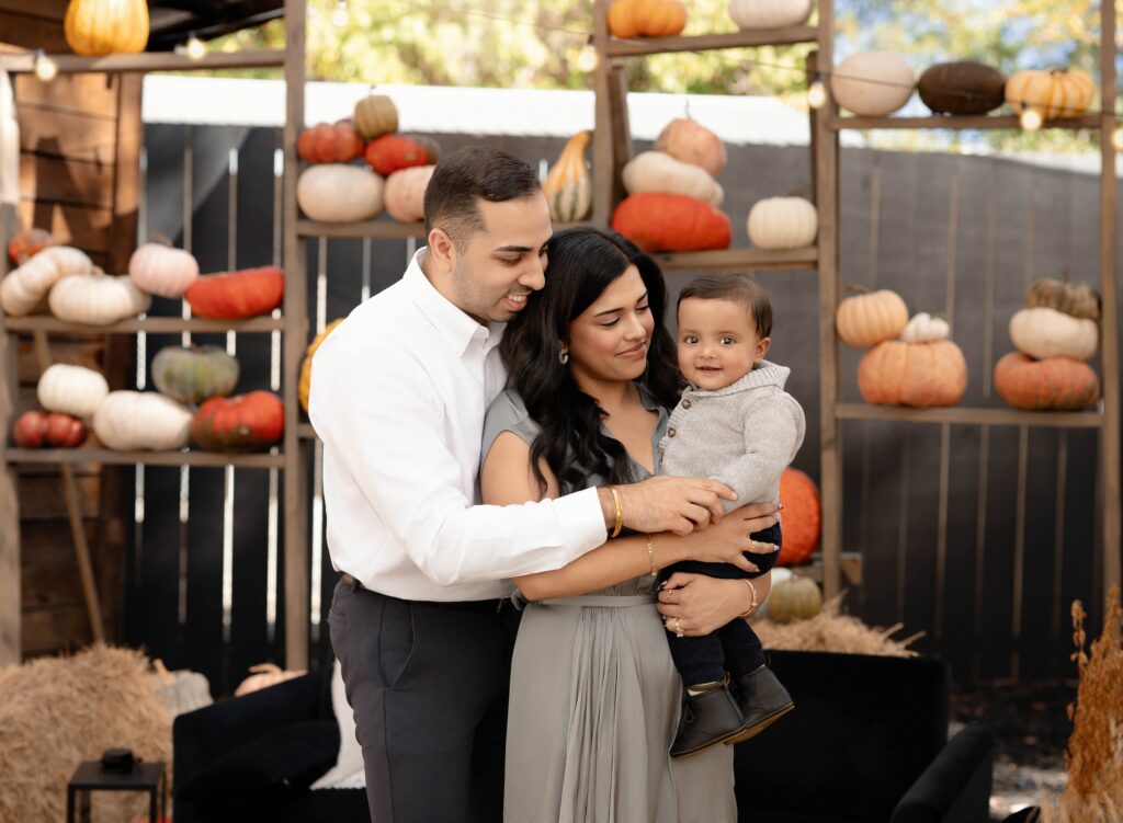 Mom and Dad smiling at baby in a setting of fall pumpkins and decor. 