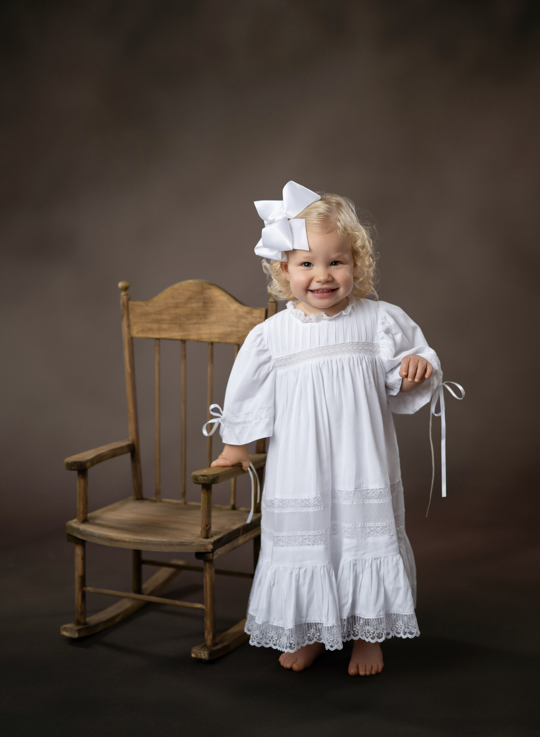 2 year old girl with white heirloom gown, standing next to a wooden rocking chair, taking portraits at Atlanta's children heirloom photography studio.