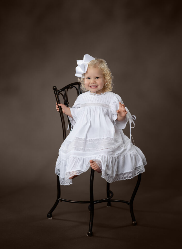 2 year old girl with white heirloom gown, sitting on a vintage chair, taking portraits at Atlanta's children heirloom photography studio.