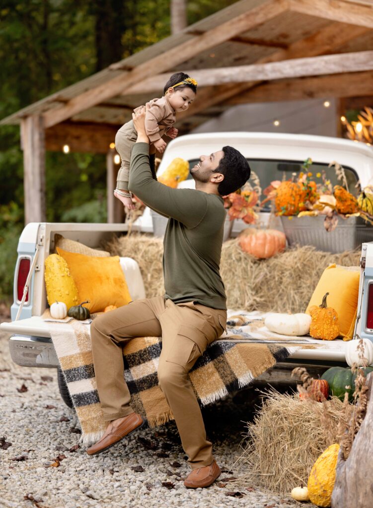Dad sitting on the back of a fall decorated vintage truck and lifting his baby in the air, smiling.
