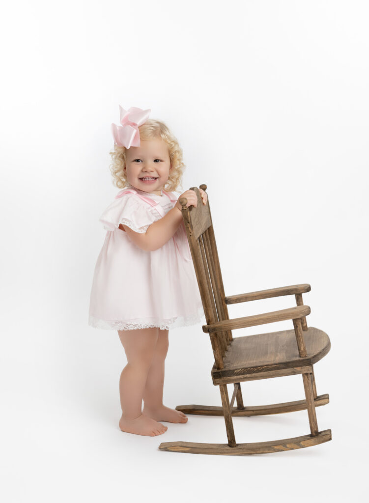 Toddler girl with pinka nd white heirloom clothing on, standing by wooden rocking chair, taking an heirloom portrait in Atlanta.