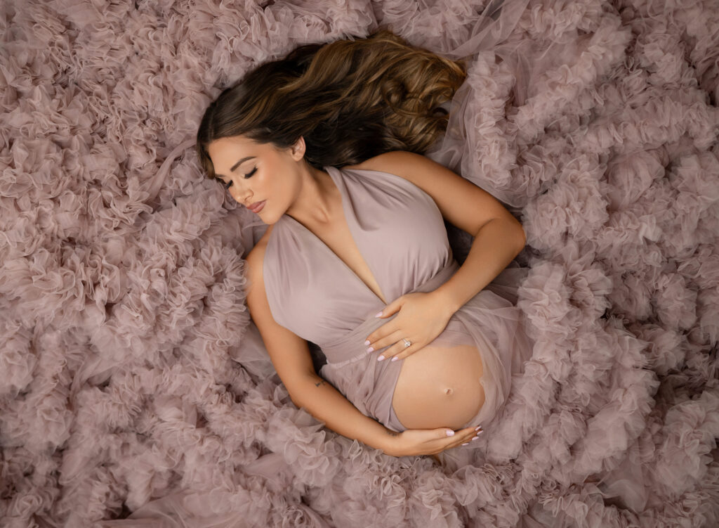 Photo of pregnant women in pink tulle gown, lying on her back and holding her baby bump. Her eyes are closed thinking of what her baby will look like.