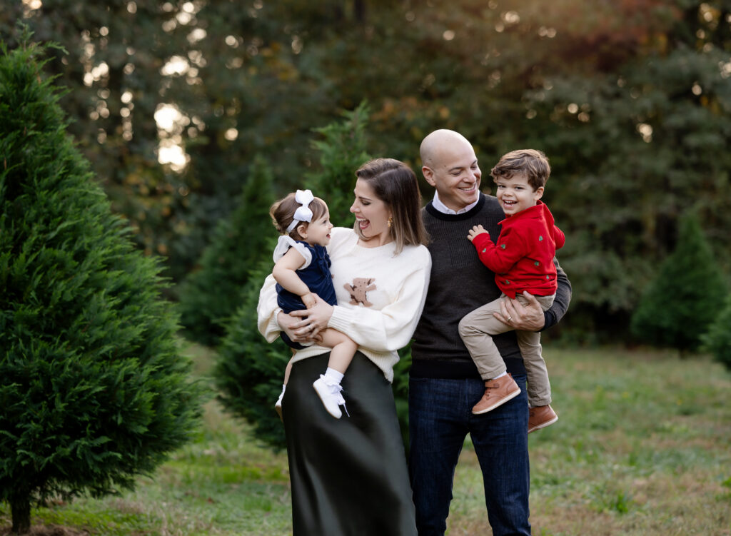 Mom and dad holding kids on their hip and laughing with them. Photographed on a tree farm for Atlanta Christmas mini sessions.