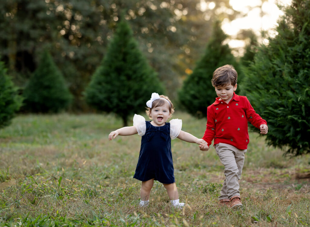 Brother and sister holding hands and running in a tree farm. Photographed by Atlanta photographer who does tree farm mini sessions.